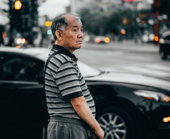 Older man appears with a loss of purpose standing on a busy street corner holding onto a cane.